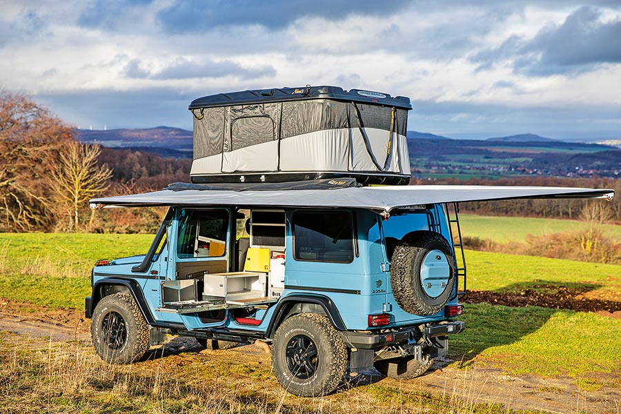 Hellblauer Mercedes G mit Camping-Ausbau, quadratischem Aufstelldach und großer Markise in weitläufiger Wiesen- und Waldlandschaft