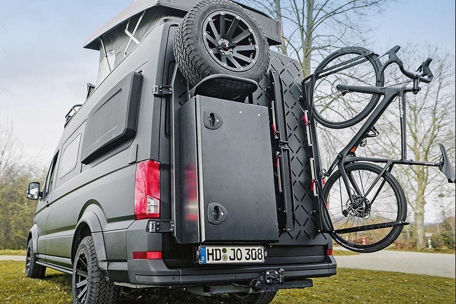 Mattschwarzer Offroad-Camper Multicamper adventure X auf MAN TGE mit Aufstelldach, Schlaf-Verbreiterungsbacken und Fahrradträger mit befestigtem Fahrrad am Heck