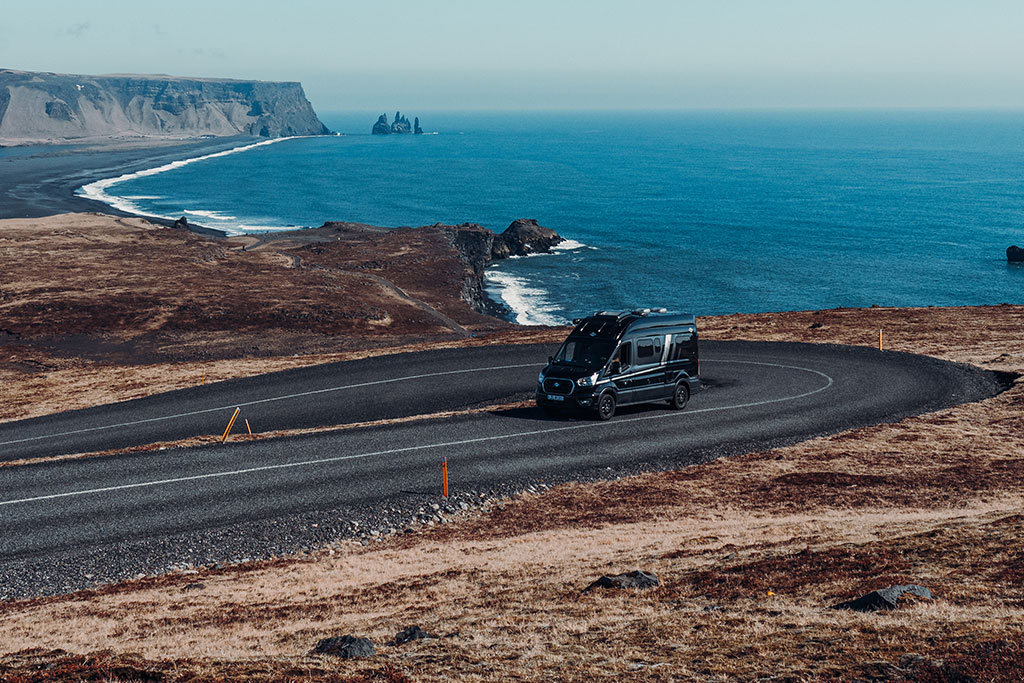 Campervan fährt auf der Straße in Island entlang einer Küste mit schwarzem Sandstrand und Felsen.