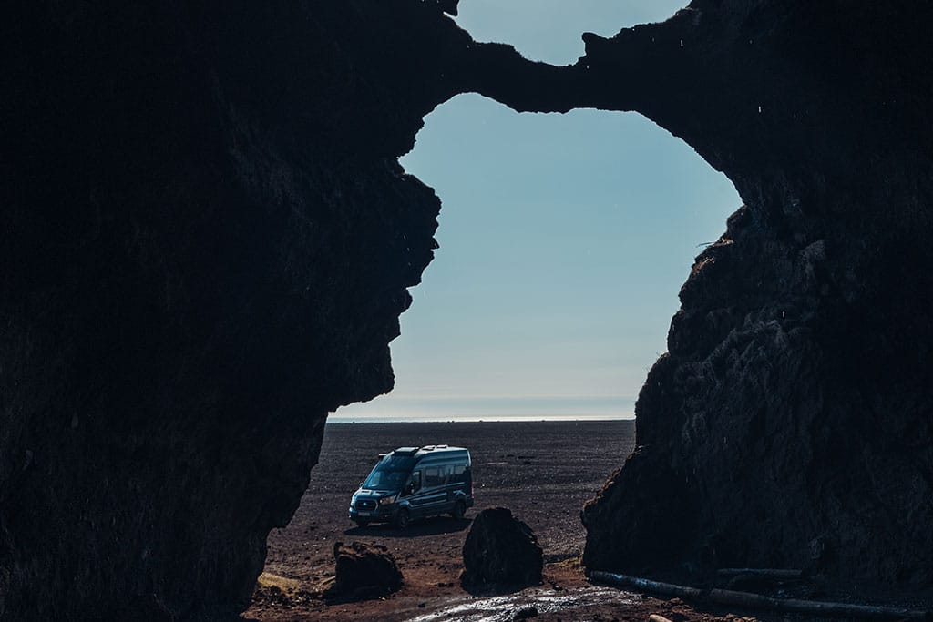 Höhle im Felsen am schwarzen Strand in Island mit Blick auf einen Campervan und das Meer.