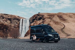 Mann sitzt vor einem CamperVan vor einem Wasserfall in Island.