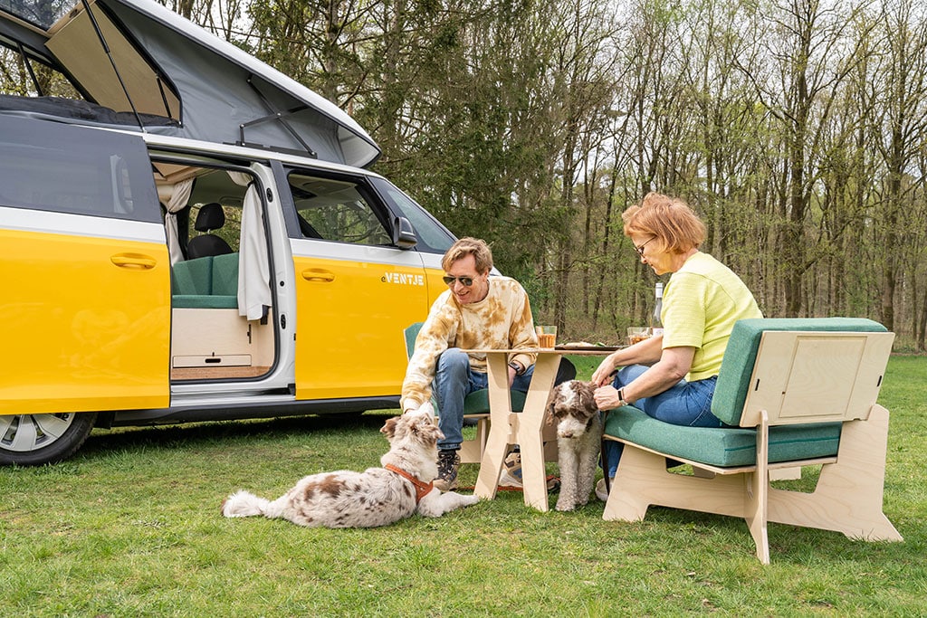 Zwei Frauen sitzen auf Holzsesseln mit türkisfarbenen Polstern an einem kleinen Holztisch, zwei Hunde sind bei ihnen, dahinter steht ein leiuchtend gelber VW ID.Buzz mit Campingausbau und aufgestelltem Schlafdach.