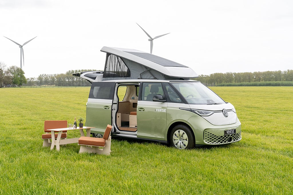 Pastellgrüner VW ID.Buzz mit Campingausbau und zwei Holzsesseln sowie einem Holztisch auf einer großen WIese, im Hintergrund einige Windräder.