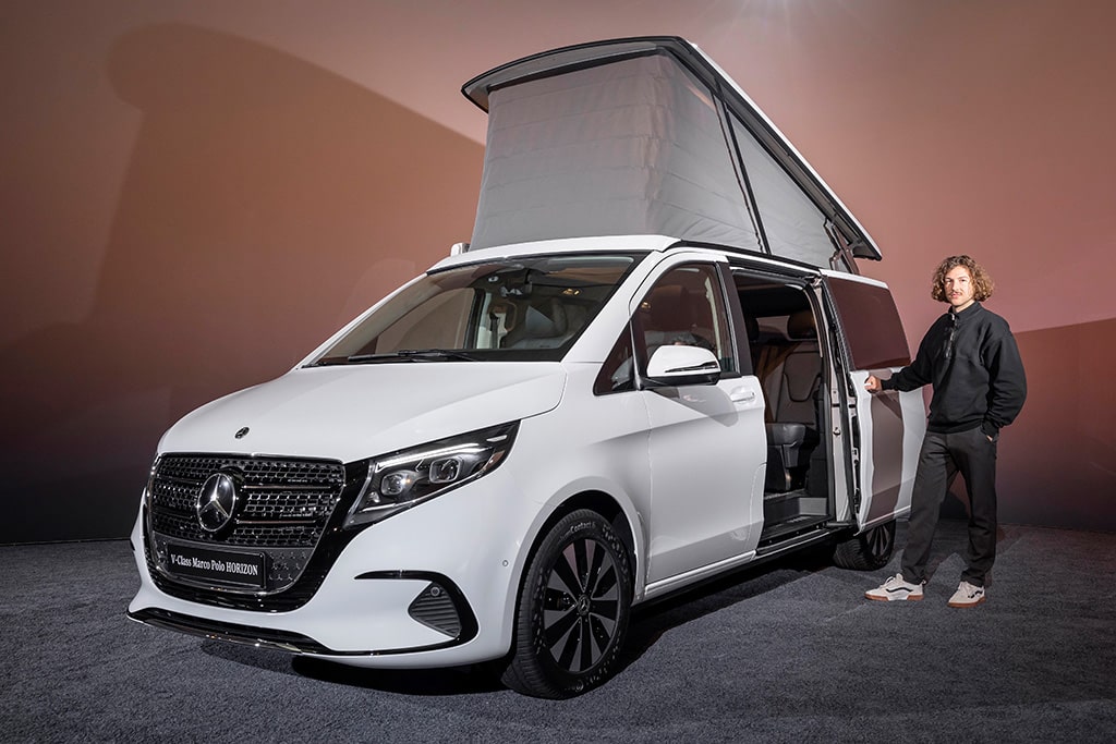 Junger Mann steht an der Schiebetür eines weißen Campingbus von Mercedes-Benz.