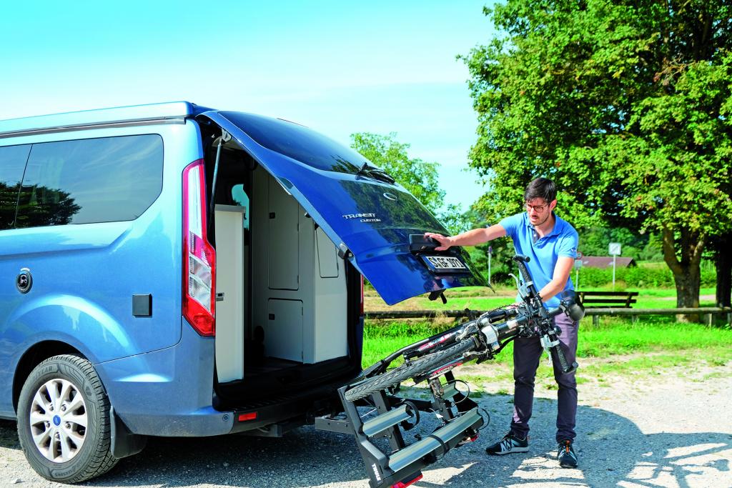 Mann klappt einen auf der Anhängekupplung eines blauen Campingbusses befestigten Fahrradträger, auf dem ein Fahrrad befestigt ist, nach unten weg, während er die Heckklappe des Fahrzeugs öffnet.