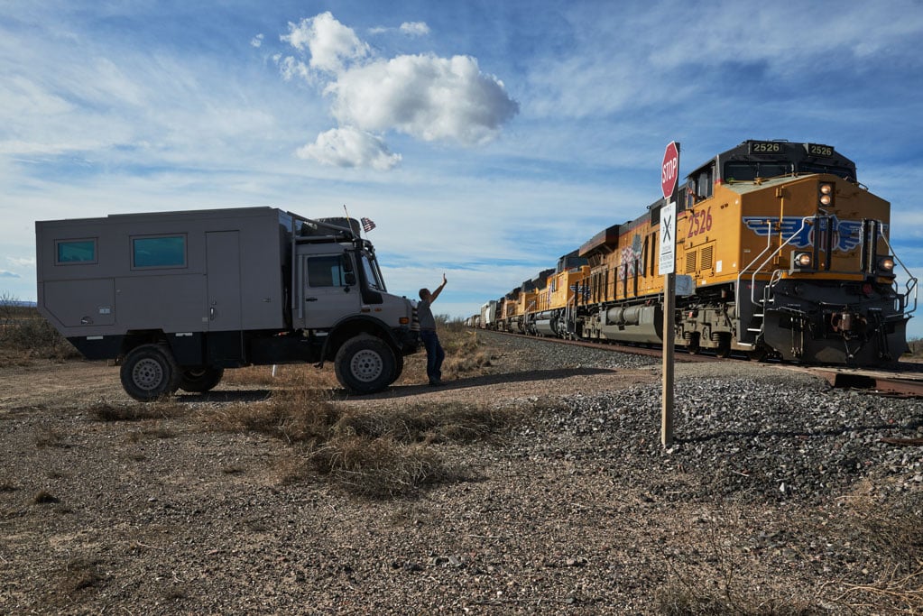 Fernreise mit dem Expeditionsmobil mogX auf Unimog: Expeditionstruck in karger Landschaft wartet vor einem Bahnübergang, während ein gelber Zug vorbeifährt.