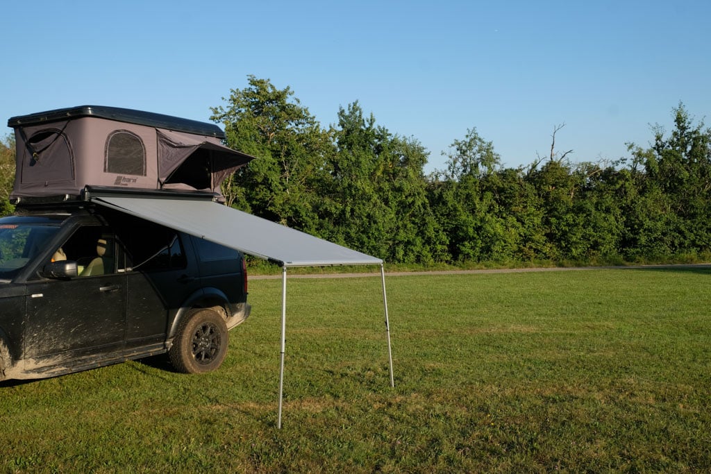 Schwarzer Geländewagen mit grauem Dachzelt und mit kleiner, an der Fahrzeugseite befestigter Markise