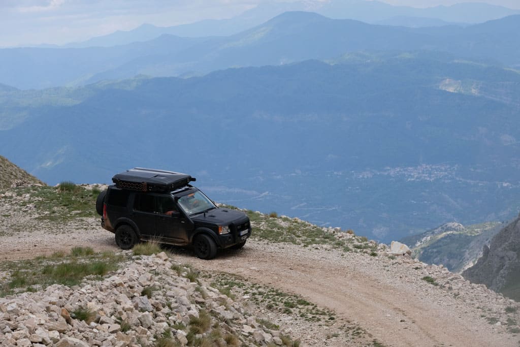 Schwarzer Geländewagen mit geschlossenem Dachzelt fährt auf einem unbefestigten Weg auf einer Bergkuppe in bergiger Landschaft